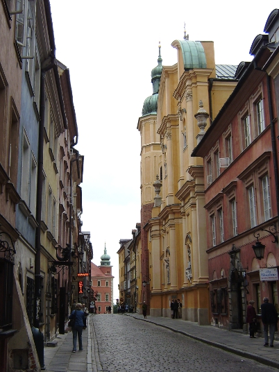 Kolorowe zdjęcie współczesne. Przedstawia ulicę i kolorowe kamienice. W oddali fragmenty Zamku Królewskiego w Warszawie. Jest to kościół augustianów na Starym Mieście w Warszawie. 