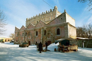 Kolorowe zdjęcie współczesne w aurze zimowej. Przedstawia synagogę w Żółkwi w 2004 r. Przed budowlą stoi samochód. Z prawej strony kram - stoisko handlowe.