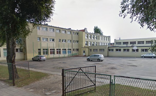 Współczesne zdjęcie. Przedstawia widok na nowoczesny budynek z wejściem, przed nim ogrodzenie i parking. Jest to V Liceum Ogólnokształcące im. Jana III Sobieskiego w Kaliszu. 