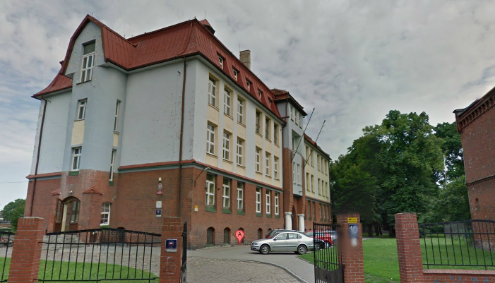 Współczesne zdjęcie. Przedstawia widok na budynek szkoły za ogrodzeniem. Jest to II Liceum Ogólnokształcącego im. Króla Jana III Sobieskiego w Grudziądzu. 