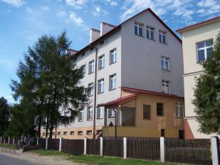 Współczesne zdjęcie. Przedstawia widok na kilkupiętrowy budynek z wejściem, przed nim ogrodzenie i droga. Jest to Szkoła Podstawowa im. Jana III Sobieskiego w Laszkach.