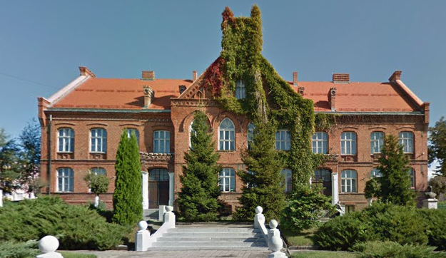 Współczesne zdjęcie. Przedstawia zabytkowy budynek z czerwonej cegły z pomarańczowym dachem. Ściany budynku przesłonięte zielonymi pędami. Przed budynkiem schody. Jest to Szkoła Podstawowa im. Jana III Sobieskiego w Gniewie. 