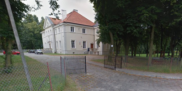 Współczesne zdjęcie. Przedstawia zabytkowy budynek za ogrodzeniem. Jest to Szkoła Podstawowa im. Jana III Sobieskiego w Woli Rasztowskiej. 