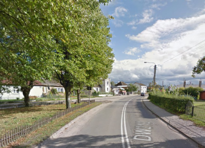 Współczesne zdjęcie. Przedstawia widok na ulicę. Z lewej strony zielone drzewa, w oddali niskie białe zabudowania. Jest to ulica Jana III Sobieskiego w Dębnicy Kaszubskiej.