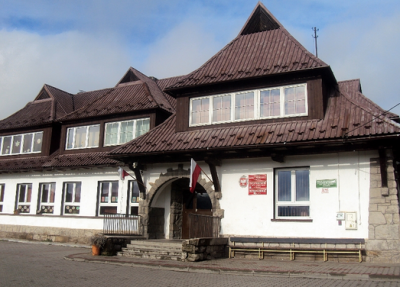 Współczesne zdjęcie. Przedstawia widok na biały, niski budynek z brązowym, strzelistym dachem. Jest to Szkoła Podstawowa im. Jana Sobieskiego w Zbludzy. 