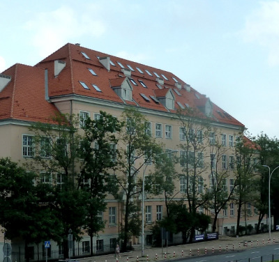 Kolorowe, współczesne zdjęcie. Przedstawia duży 
budynek z pomarańczowym dachem. Jego fasada ukryta jest za drzewami. Jest to Liceum im. Jana III Sobieskiego w Warszawie. 