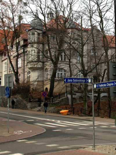 Zdjęcie współczesne, kolorowe. Na pierwszym planie skrzyżowanie dwóch ulic i drogowskazy z nazwami: Jana Sobieskiego i Fryderyka Chopina. W tle kamienice przesłonięte drzewami.