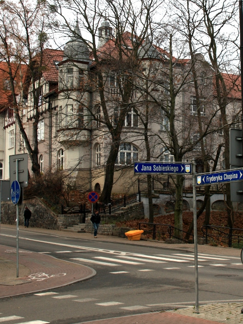 Zdjęcie współczesne, kolorowe. Na pierwszym planie skrzyżowanie dwóch ulic i drogowskazy z nazwami: Jana Sobieskiego i Fryderyka Chopina. W tle kamienice przesłonięte drzewami.