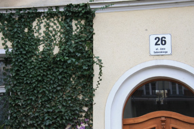 Zdjęcie współczesne, kolorowe. Po prawej stronie fragment wejścia, nad nim napis: 26 ul. Jana Sobieskiego. Po lewej okno przesłonięte zieloną roślinnością.