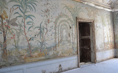 Na zdjęciu widzimy ścianę z wejściem. Na ścianie fresk a na nim drzewka z owocami i palmy. Za roślinami zarys budowli. Kolory delikatne, monochromatyczne.