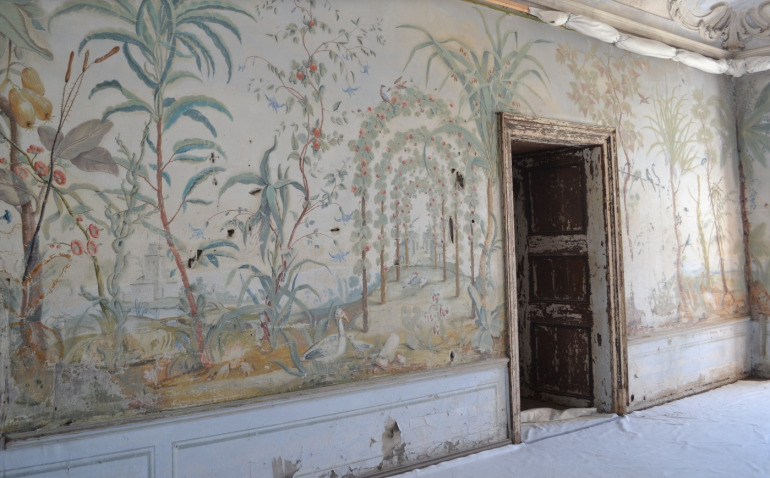 Na zdjęciu widzimy ścianę z wejściem. Na ścianie fresk a na nim drzewka z owocami i palmy. Za roślinami zarys budowli. Kolory delikatne, monochromatyczne.