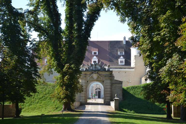 Zdjęcie przedstawia drogę prowadzącą do zabytkowej bramy za którą widzimy budowlę. Przy drodze rosną wysokie drzewa.  