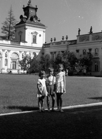 Na dziedzińcu pałacu stoi troje dzieci. Stoją od najwyższego do najniższego, obejmują się ramionami. Za nimi fragment pałacu.