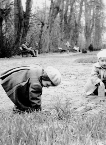 Park, ujęcie z poziomu dziecka. Dwoje dzieci kuca na trawie. Ubrane w berety i płaszczyki. W tle ludzie siedzą na ławkach albo spacerują.