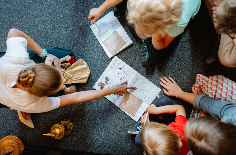 Widok z góry na podłogę na której siedzą małe dzieci. Przed nimi kartki z zadaniami edukacyjnymi. Kobieta siedząca po lewej stronie wskazuje swoją ręką na zadanie na kartce.