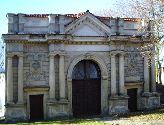 Kolorowe zdjęcie współczesne. Przedstawia niską budowlę architektoniczną. Jest to brama z dużym wejściem pośrodku oraz dwoma mniejszymi po bokach. Na fasadzie kolumny. Jest to brama zamku w Białej Podlaskiej. 