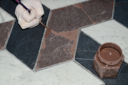 Kolorowe zdjęcie na którym widzimy zniszczony fragment posadzki. Po prawej stronie słoik z farbą, po lewej stronie ręka w białej rękawiczce z pędzlem. Jest to retusz uzupełnień posadzki w Bibliotece Króla.