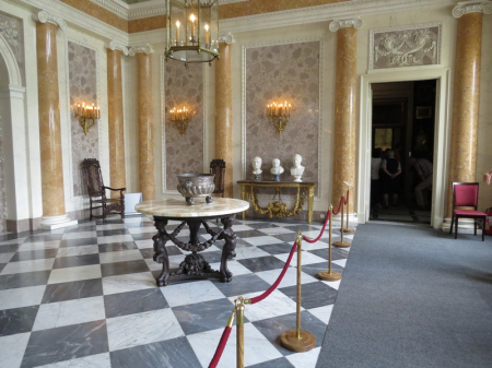 Kolorowe zdjęcie współczesne. Przedstawia salę pałacową z czarno-białą posadzką na której stoi stół ze srebrną wazą. W głębi, na stole trzy białe popiersi męskie.  Po prawej stronie wejście do sali.