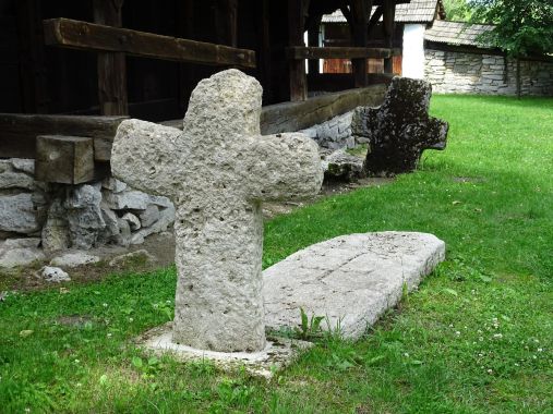 Kolorowe zdjęcie współczesne. Przedstawia schematyczny nagrobek kamienny z krzyżem. Płaska płyta leży na trawie. Na drugim planie pociemniały od mchu krzyż i zabudowania.
