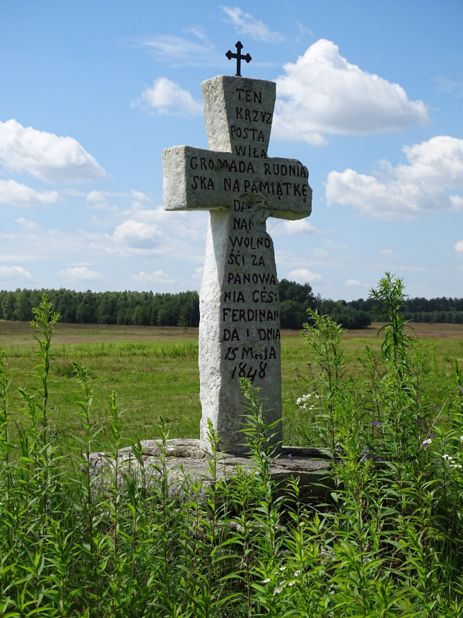Kolorowe zdjęcie współczesne. Przedstawia samotnie stojący krzyż na tle zielonego pejzażu. Na ramionach krzyża napisy. 
