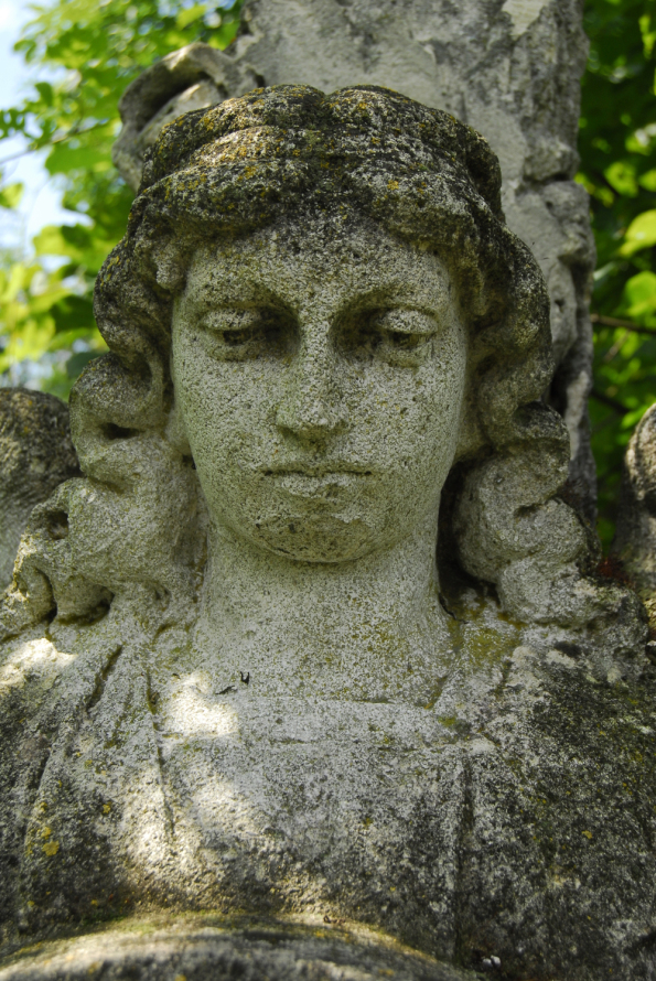 Kolorowe zdjęcie współczesne. Przedstawia starą, kamienną rzeźbę nagrobną. Jest to portret kobiety z długimi włosami, która spuszcza oczy w dół. 