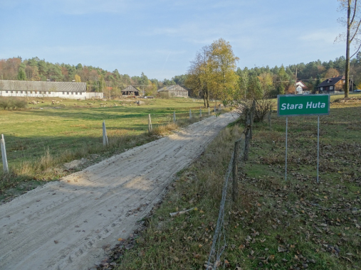 Kolorowa fotografia współczesna. Przedstawia drogę wiejską i napis "Stara Huta". Widzimy zabudowania, drewniane stodoły i domy. 