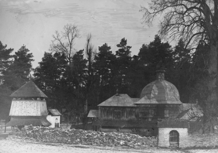 Czarno biała fotografia. Przedstawia stary, drewniany kościół i dzwonnicę. Przed kościołem ogrodzenie murowane z kamienia i bramka wejściowa. Za kościołem las. 