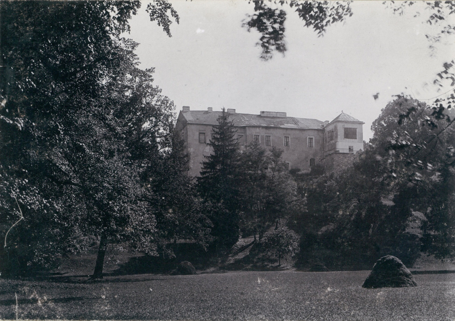 Czarno-białe, archiwalne zdjęcie. Na pierwszym planie ścięty trawnik i kopka trawy. Powyżej wzgórze z drzewami i krzewami. Na podniesieniu stary budynek, który wygląda na pałac. Po prawej stronie zdjęcia duże drzewa liściaste. 