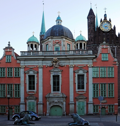 Fotografia kolorowa, współczesna. Przedstawia fasadę kościoła Wniebowzięcia NMP w Gdańsku, w której znajduje się kaplica Królewska. Fasada pomarańczowa, z jasno-zielonymi oknami i okiennicami. Portal główny i dwa portale boczne mają zielone drzwi. Całość w stylu barokowym.