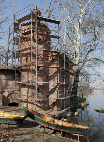 Budynek pompowni przypominający ceglany niewielki zamek otoczony rusztowaniami.