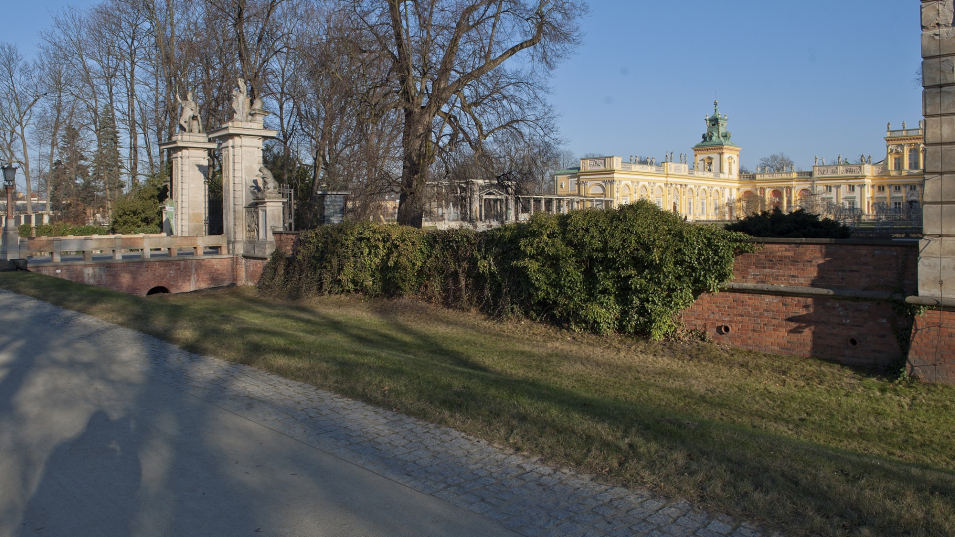 Mur porośnięty bluszczem, obok brama główna, w tle pałac wilanowski.